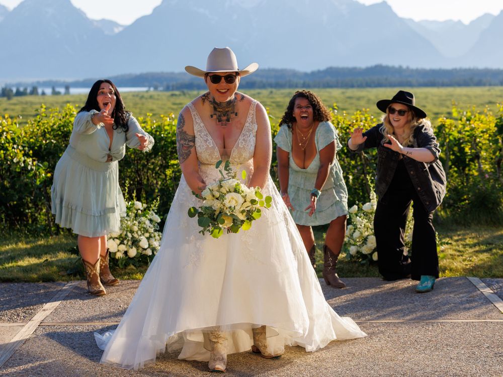 A bridal party walks and laughs together on a path through a vineyard with snow-capped mountains in the background.
