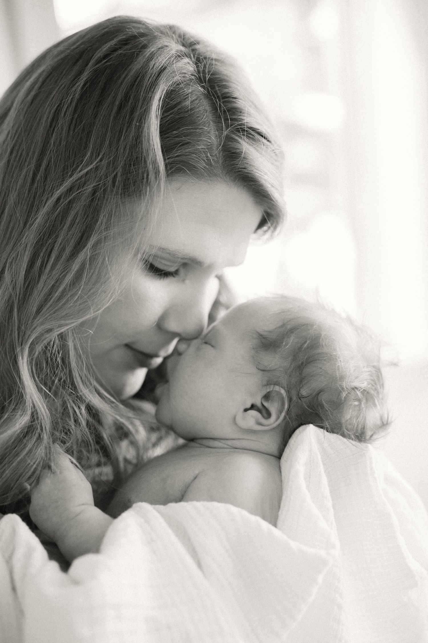 A tender moment captured in black and white as a mother snuggles close with her sleeping newborn baby wrapped in soft fabric.