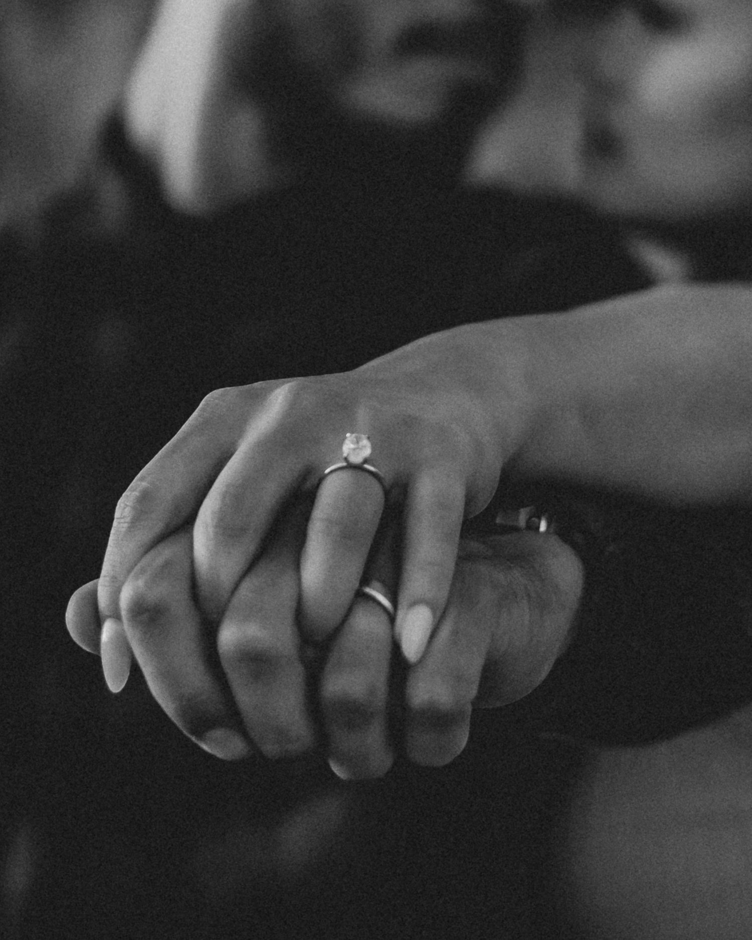 Close-up black and white photo showing two hands clasped together with an engagement ring on display.