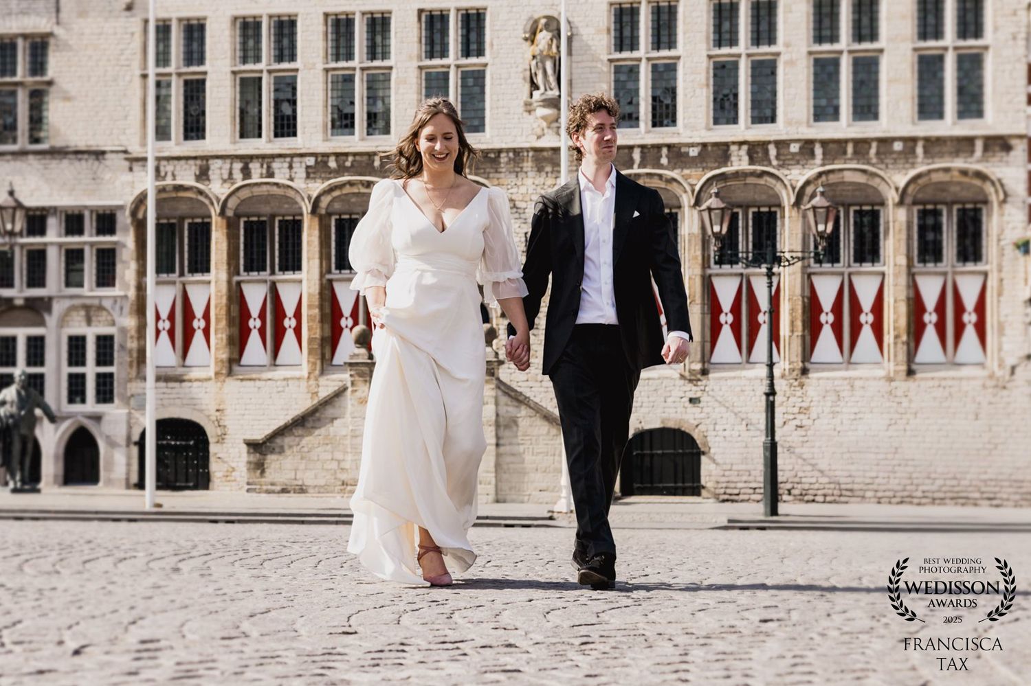 A couple in wedding attire walking hand in hand on a plaza in front of a historic Gothic building with ornate architecture.