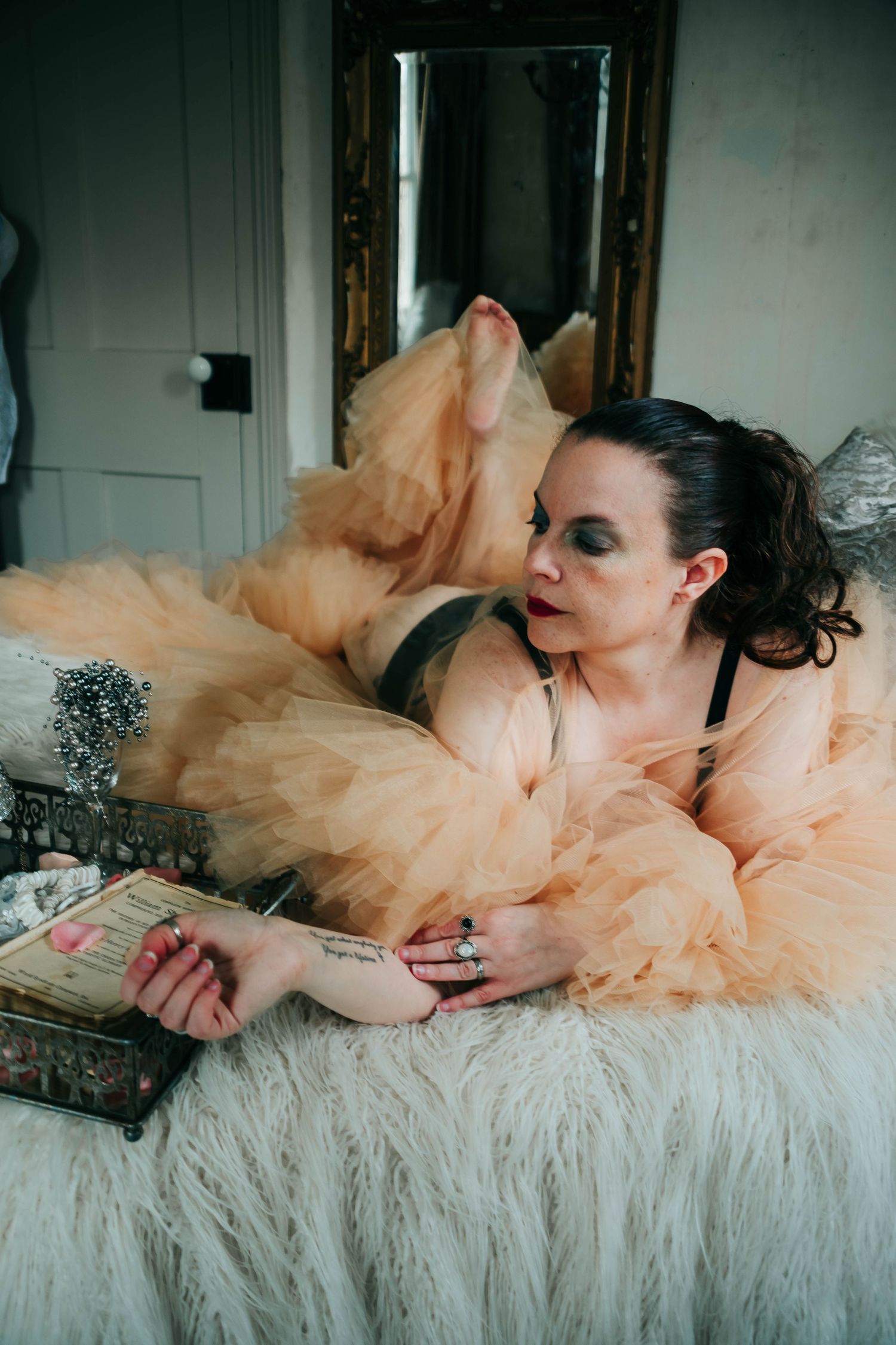 Figure in a peach tulle gown reclines on white faux fur while reading a vintage book in moody boudoir setting.
