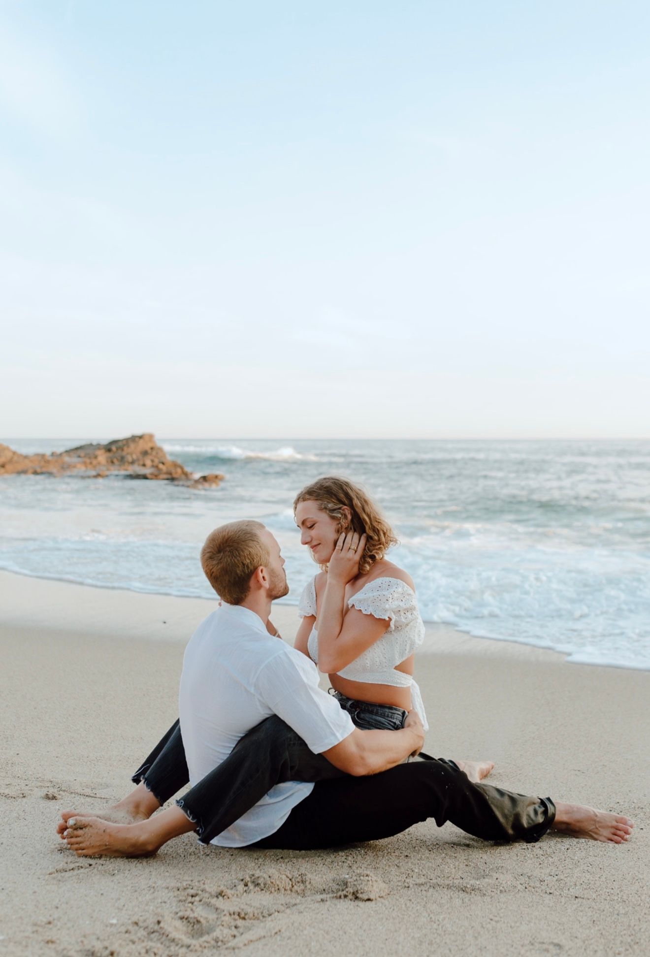 Couple sits intimately together on a sandy beach with crashing waves and rocky coastline in the background.