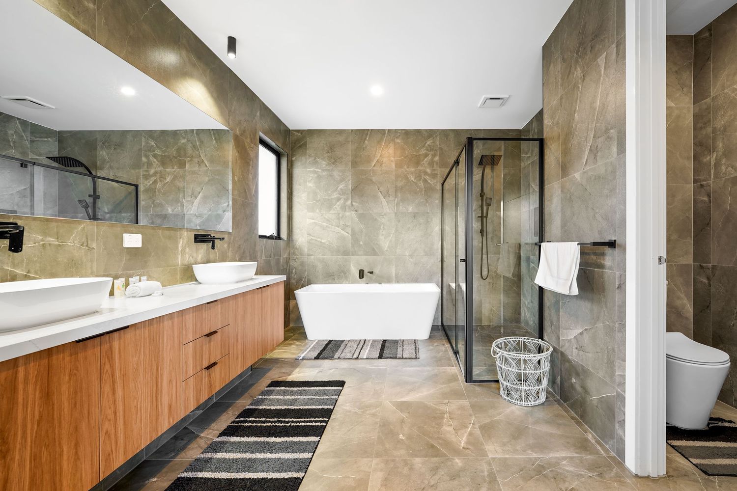 Modern luxury bathroom with freestanding tub, wood vanity, glass shower and beige marble tile walls and floors.