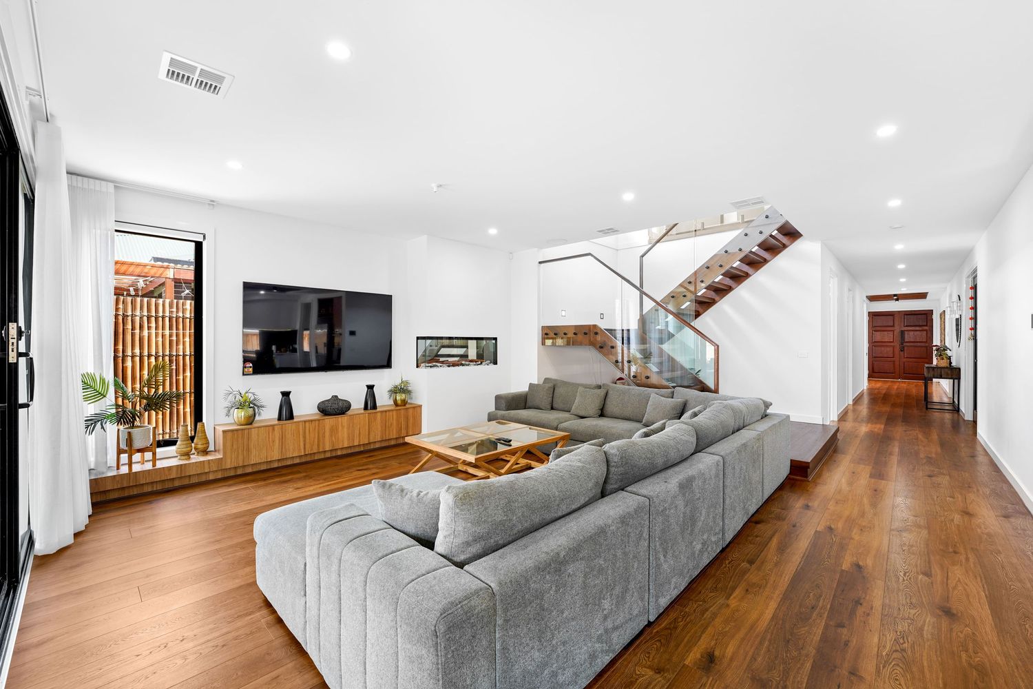 Modern open concept living room with grey sectional sofa, wall mounted TV, and hardwood floors leading to staircase.