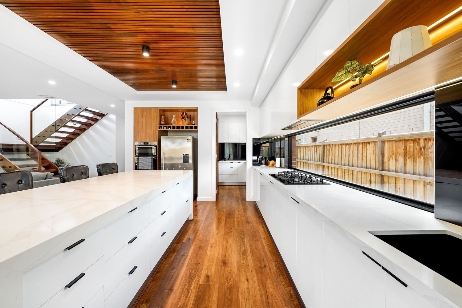 Modern kitchen interior with white countertops, hardwood flooring and wood ceiling featuring sleek cabinetry and open design.