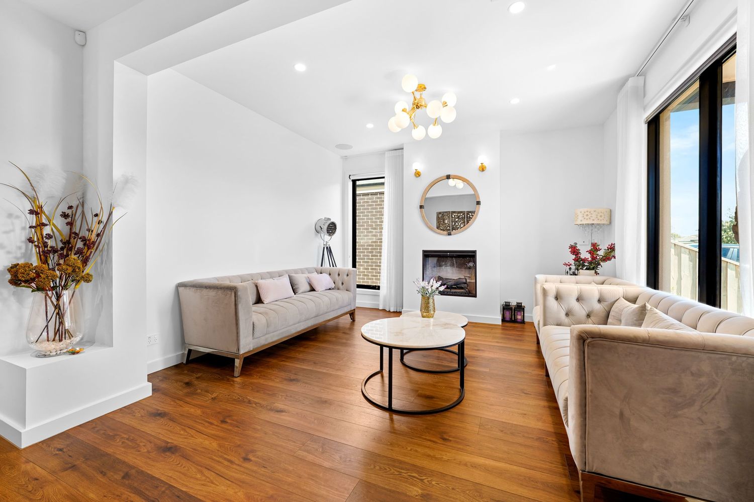 Modern living room with beige tufted sofas, hardwood floors, and contemporary lighting fixture in an all-white space.