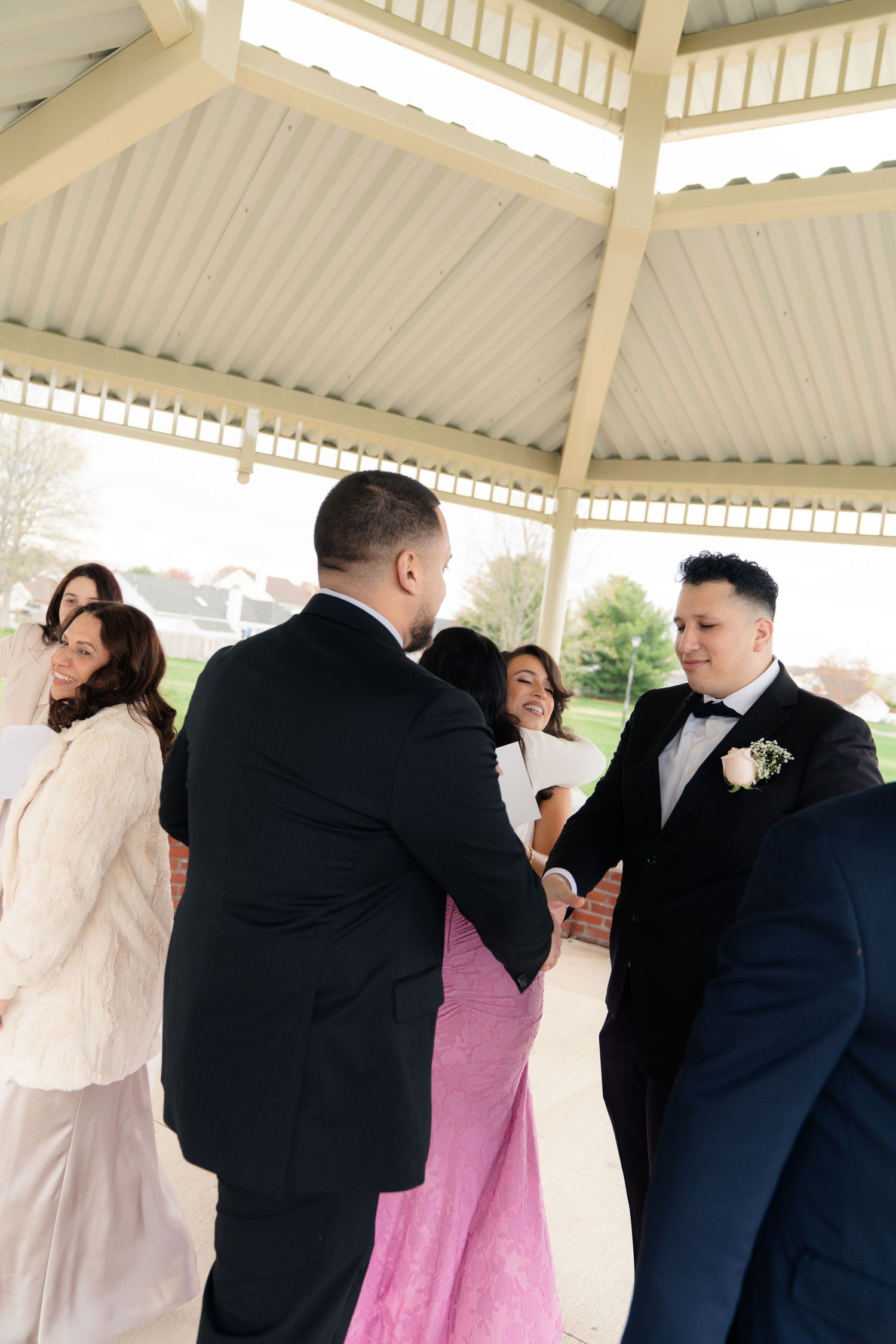 People gather under a white gazebo at a formal event wearing elegant attire including tuxedos and formal dresses.