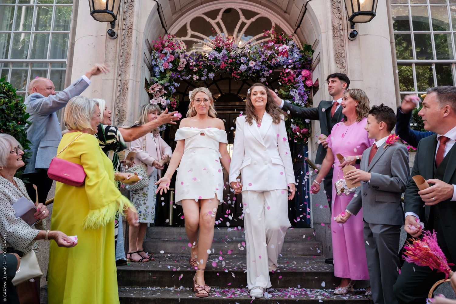 Wedding guests celebrate and dance together outside a grand building with twinkling lights and floral decorations.