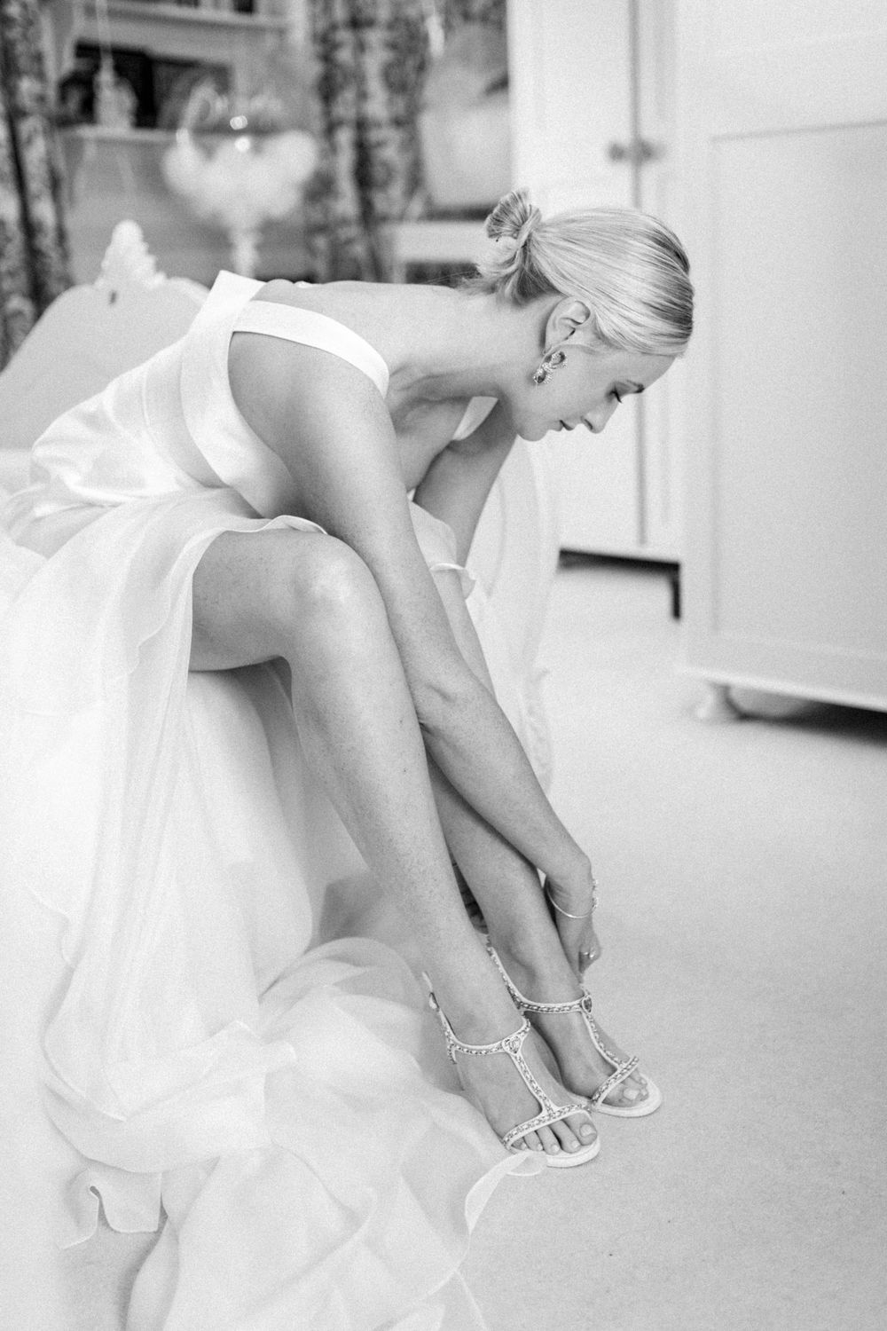 Bride in white dress sits while fastening elegant strappy sandals in a candid black and white moment.