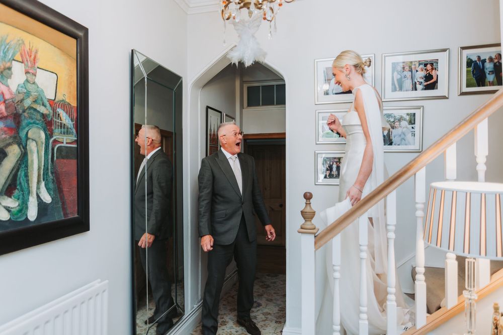 Wedding party members walk through an elegant white hallway with stairs and framed photos on the wall.