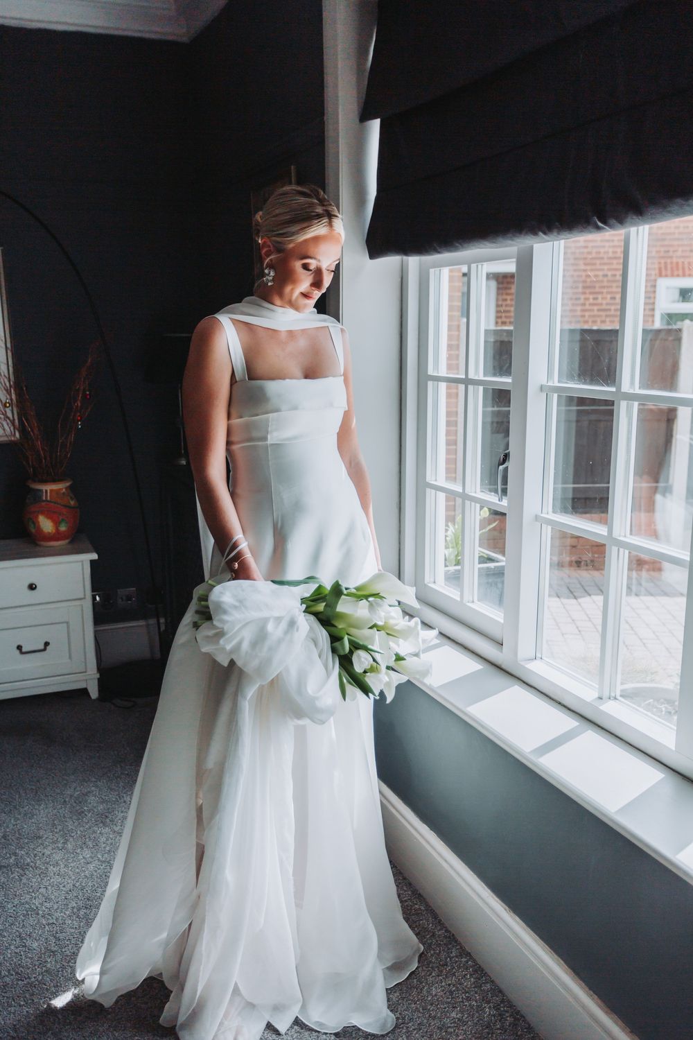 A bride in a flowing white wedding dress poses next to large windows in a room with natural lighting.