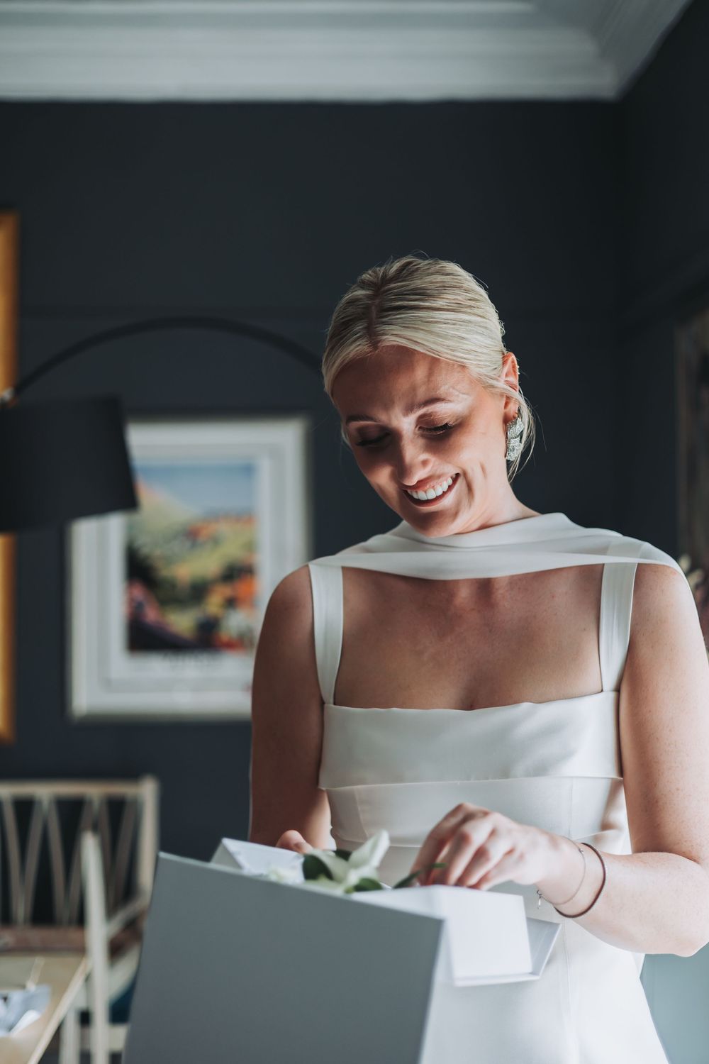 Bride in elegant white sleeveless dress smiles while looking down at a gift box in an indoor setting.