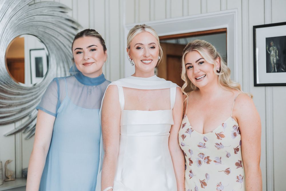 Three bridesmaids pose together wearing soft blue, white, and floral dresses in a light grey interior setting.