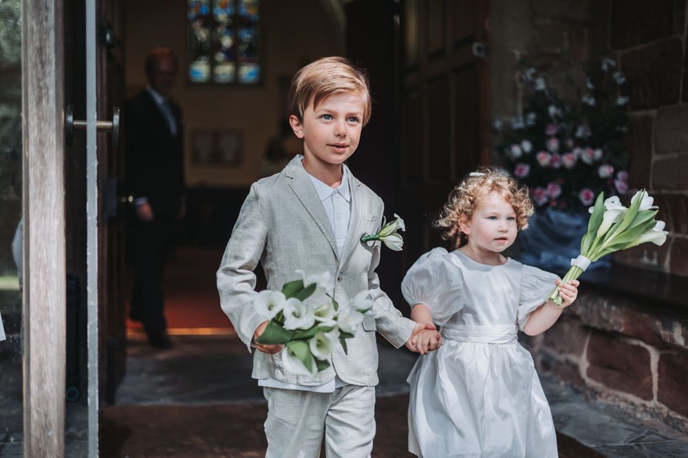 A wedding party carries white floral bouquets while walking down a brick-lined pathway outside a church.