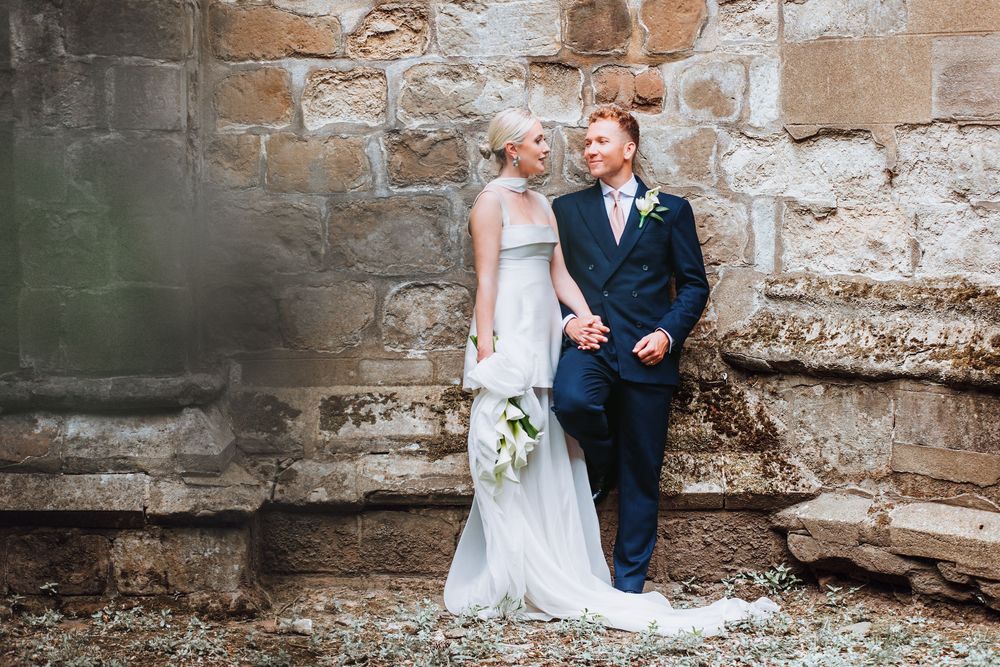 Newlywed couple poses romantically against rustic stone wall during their wedding day.