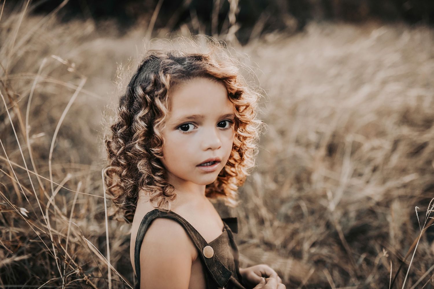 a curly haired little girl stands in a field at sunset