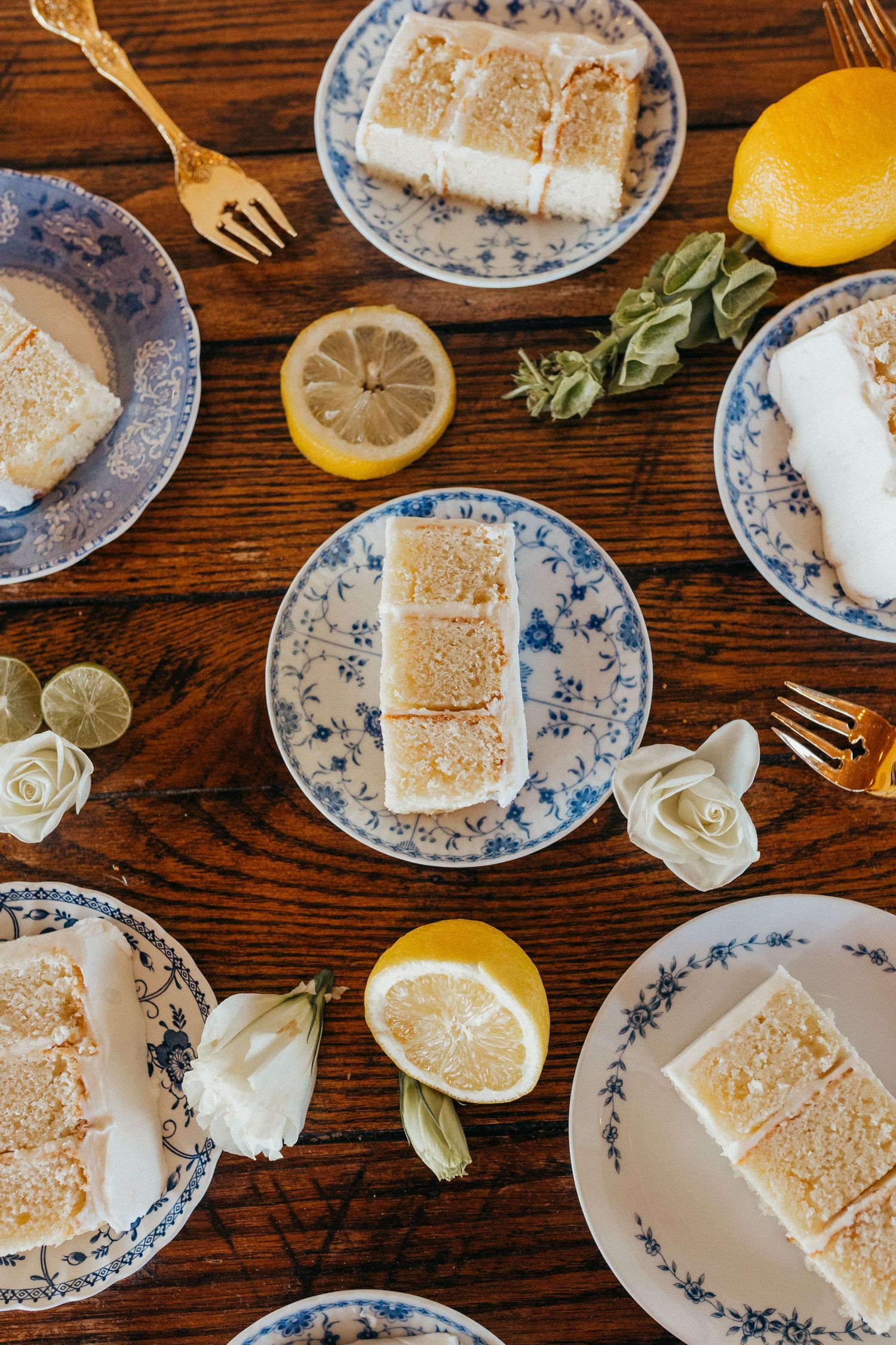 Slices of lemon cake on blue and white china plates with fresh citrus and roses on a wooden table.
