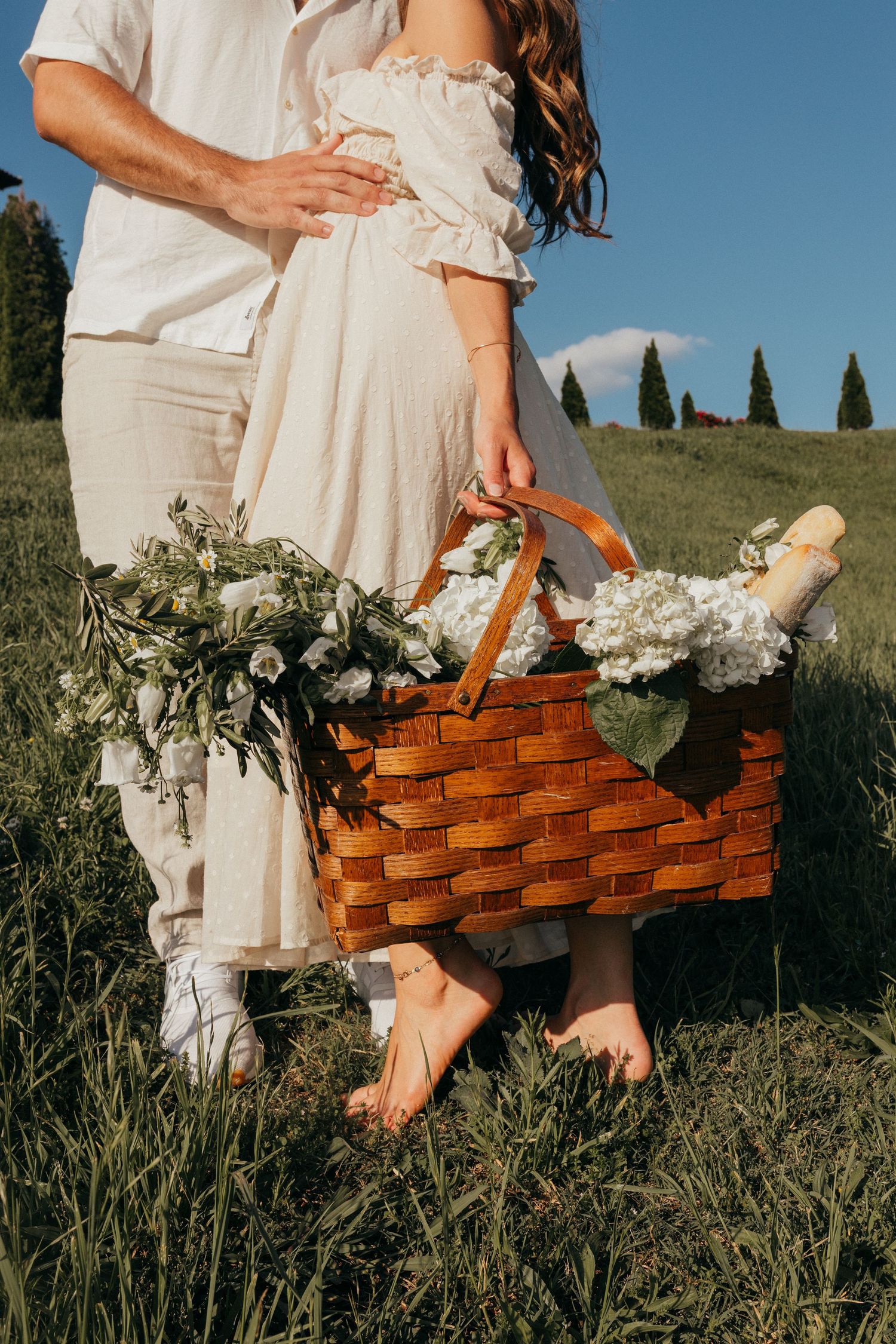 A couple in white clothing holds a wooden picnic basket filled with flowers against a pastoral backdrop of cypress trees.