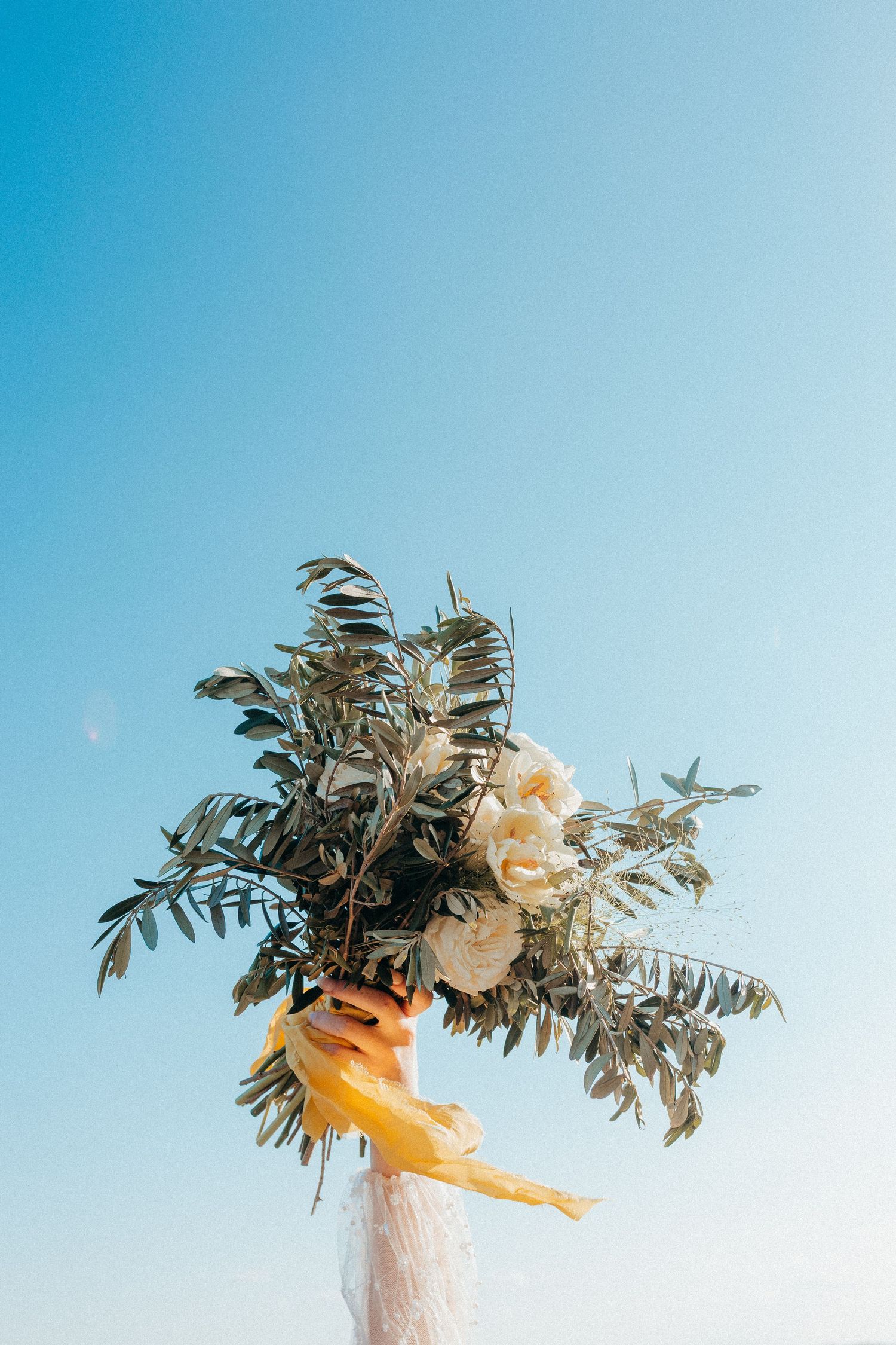 Hand holding up a beautiful floral bouquet with white flowers and greenery against a bright blue sky.