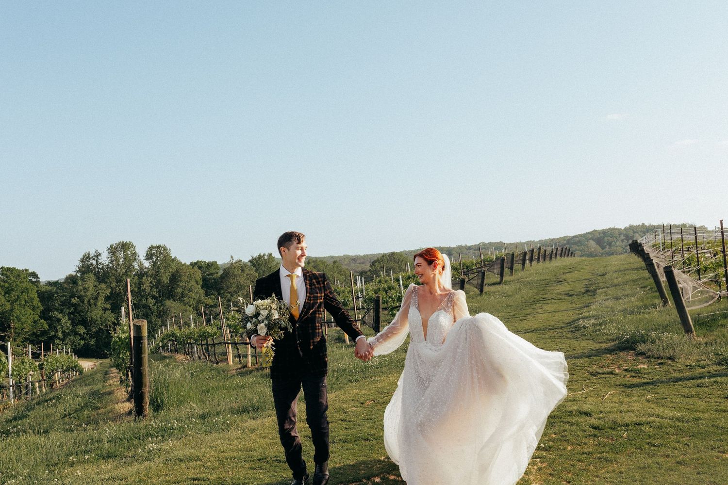 A couple holding hands while running through a vineyard with hills and vines stretching into the distance on a sunny day.