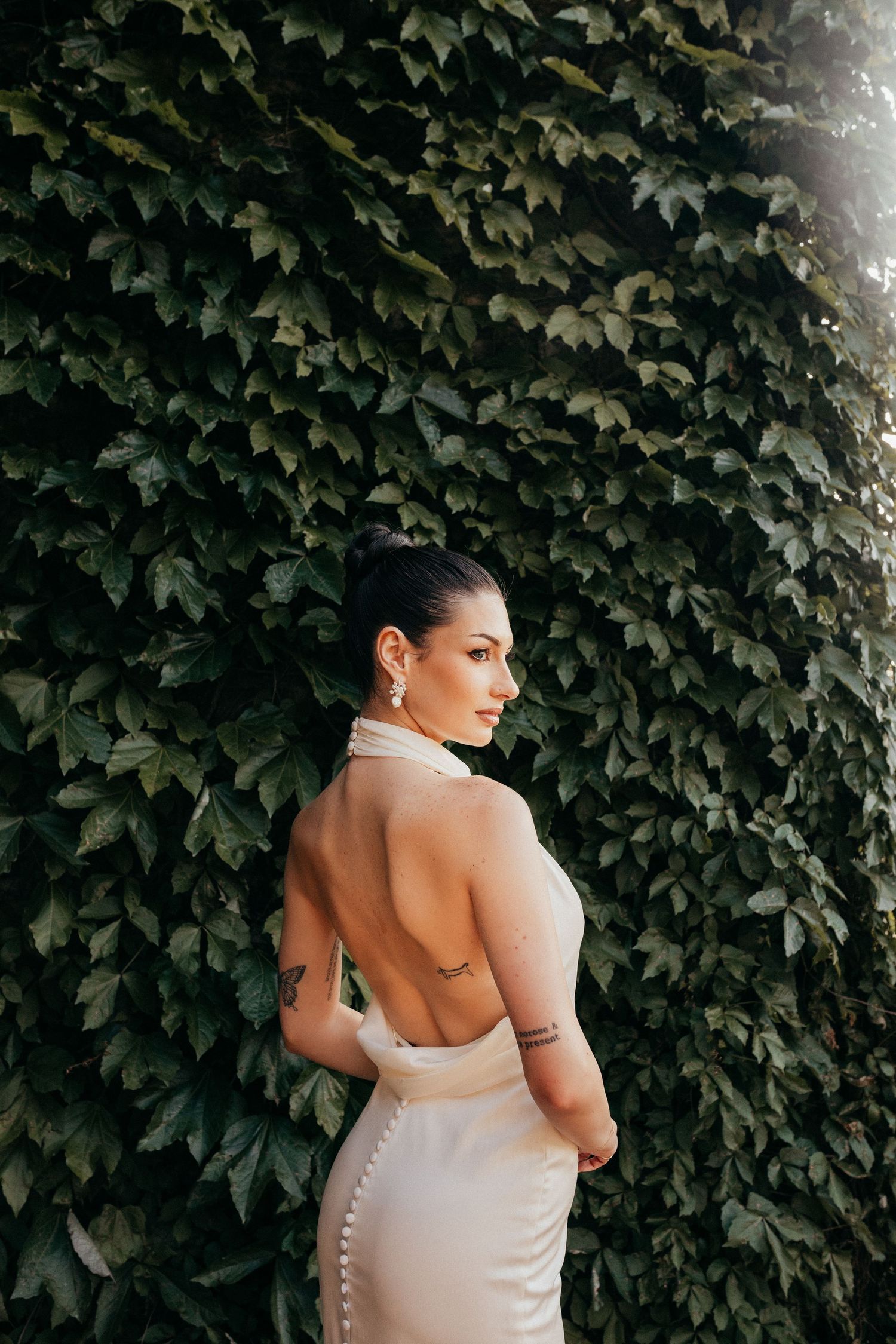 Elegant white backless dress with button details photographed against an ivy-covered wall in natural lighting.