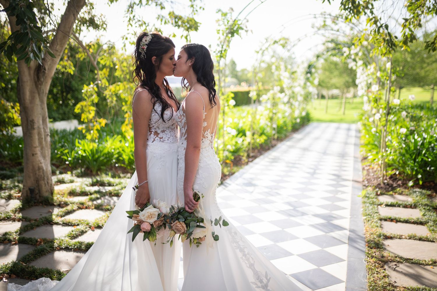 Wedding couple in matching white dresses share moment on garden path.