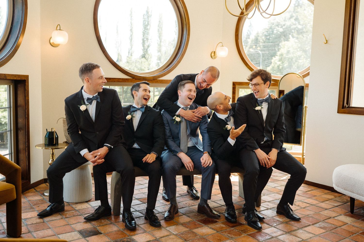 Groomsmen in black tuxedos share a lighthearted moment while seated together in an elegant room with round windows.