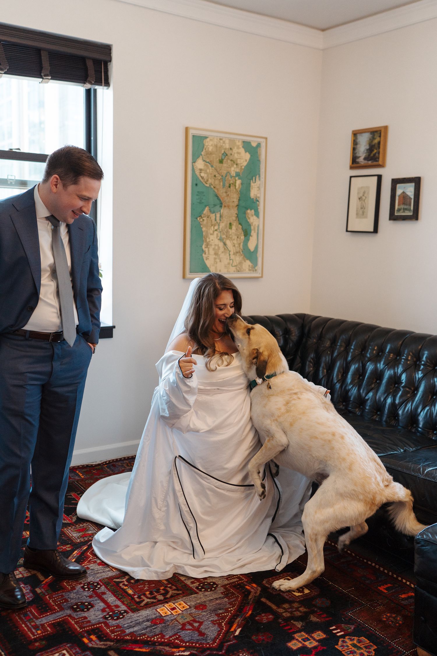 Dog greets bride in white wedding dress while suited figure looks on in cozy room with vintage decor and framed map.
