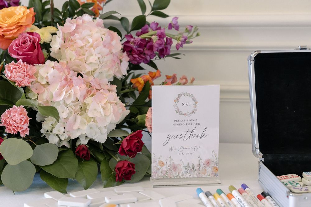 A floral arrangement with pink peonies and eucalyptus sits next to a guestbook card and writing supplies on a white table.