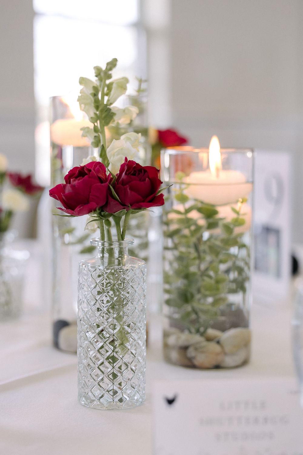 Crystal vases with red roses and greenery arranged on a white table with glowing candles in glass holders.