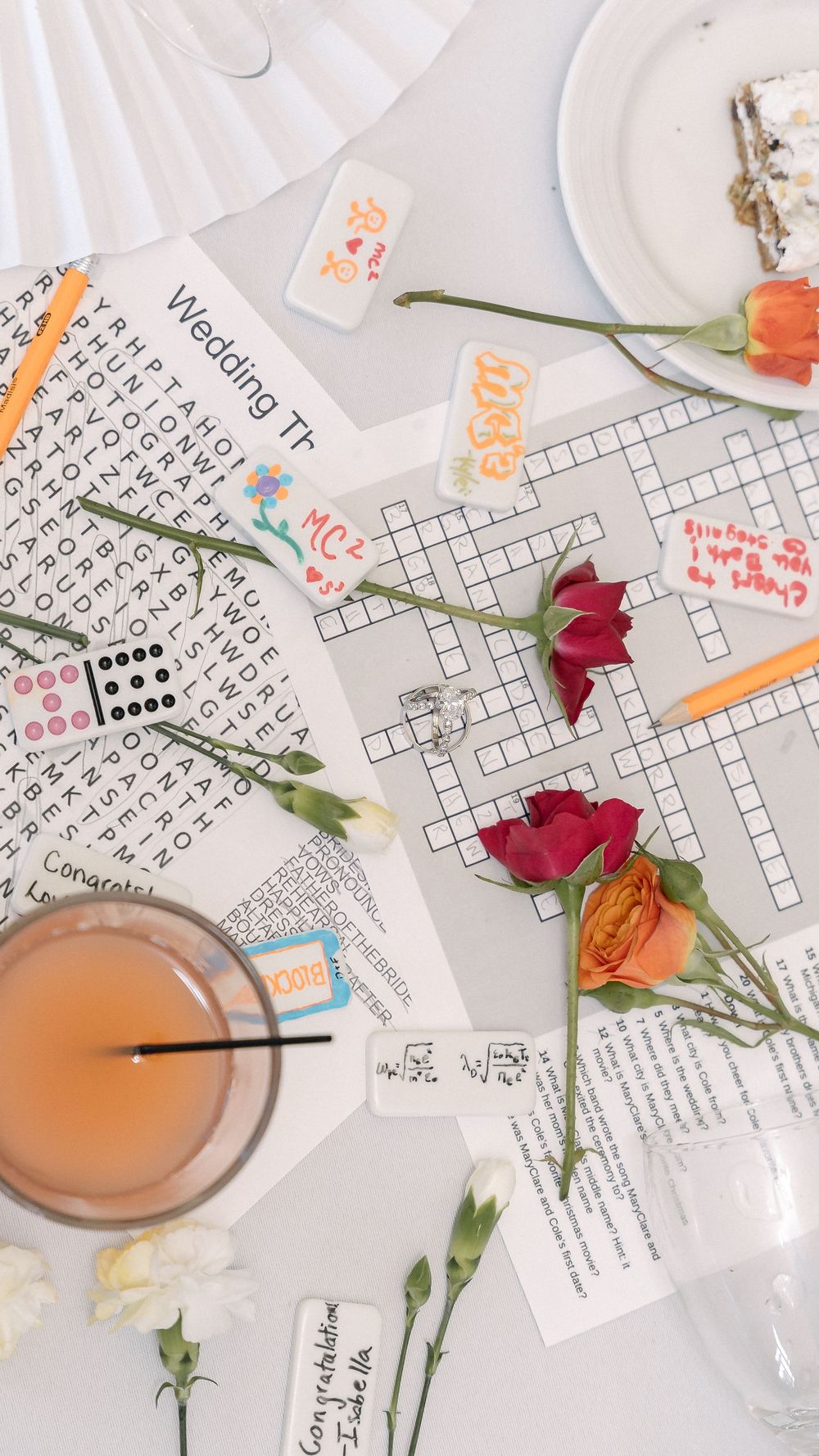 Top view of crossword puzzles with orange drink, scattered roses, and dice pieces on white table.