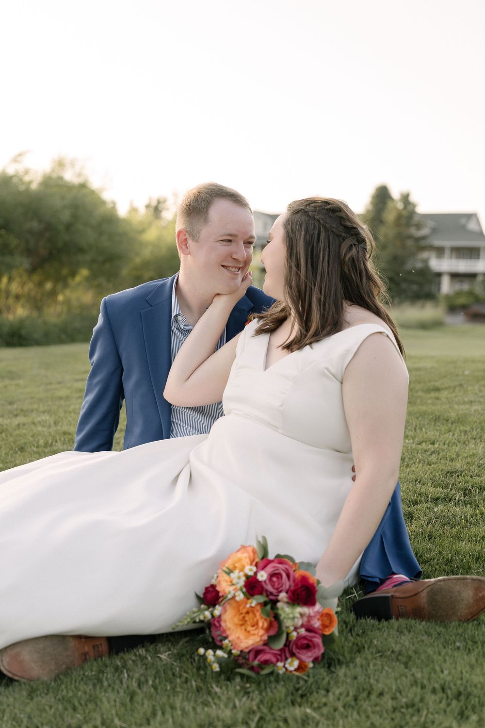 Wedding couple sitting on grass sharing an intimate moment with a colorful bouquet nearby at sunset.