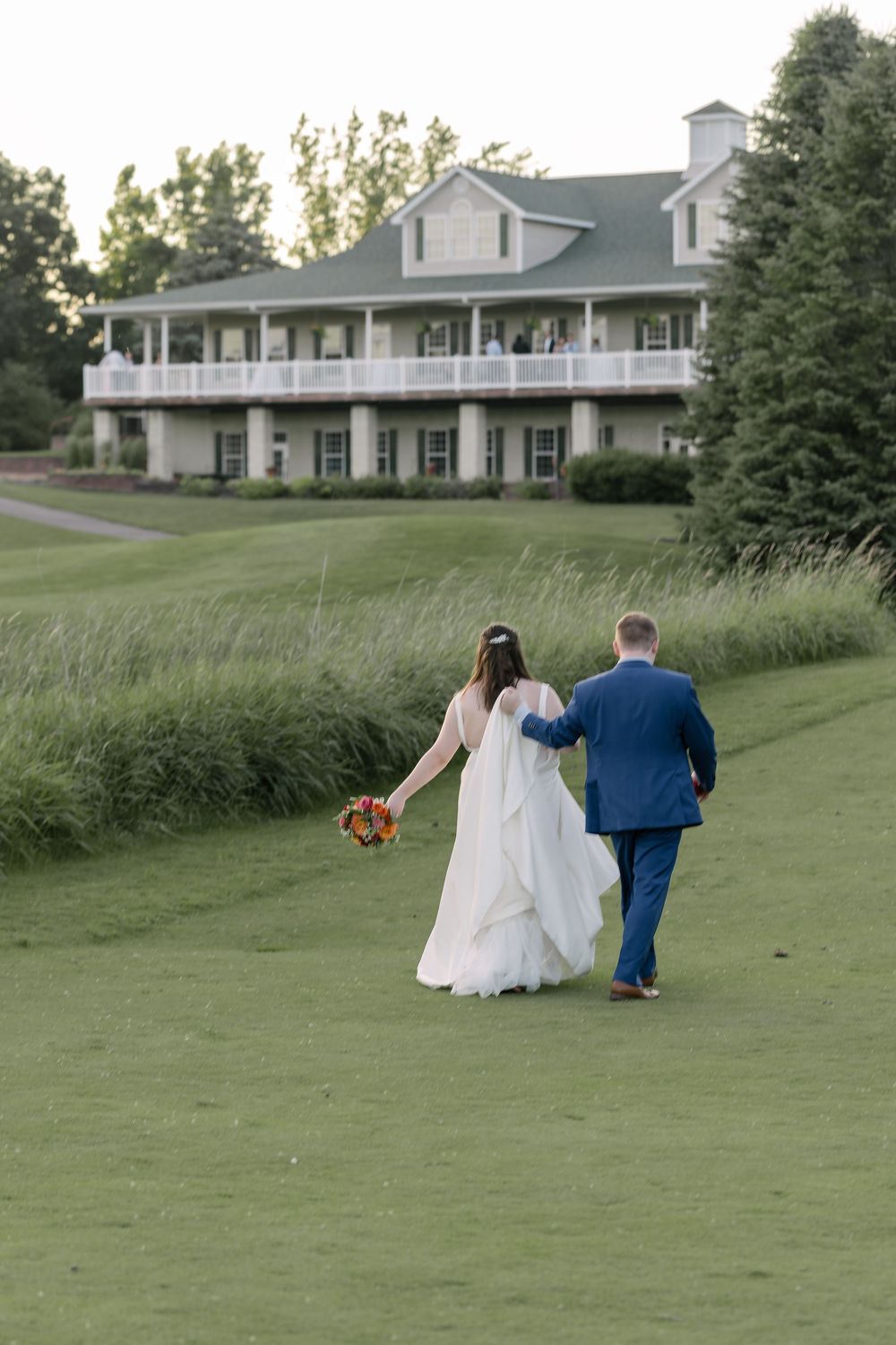 Couple walks hand in hand across a rolling green lawn with white clubhouse building visible in background.