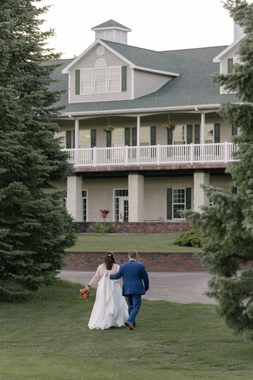A couple walks together on a lush lawn in front of an elegant white colonial-style venue with a wraparound porch.