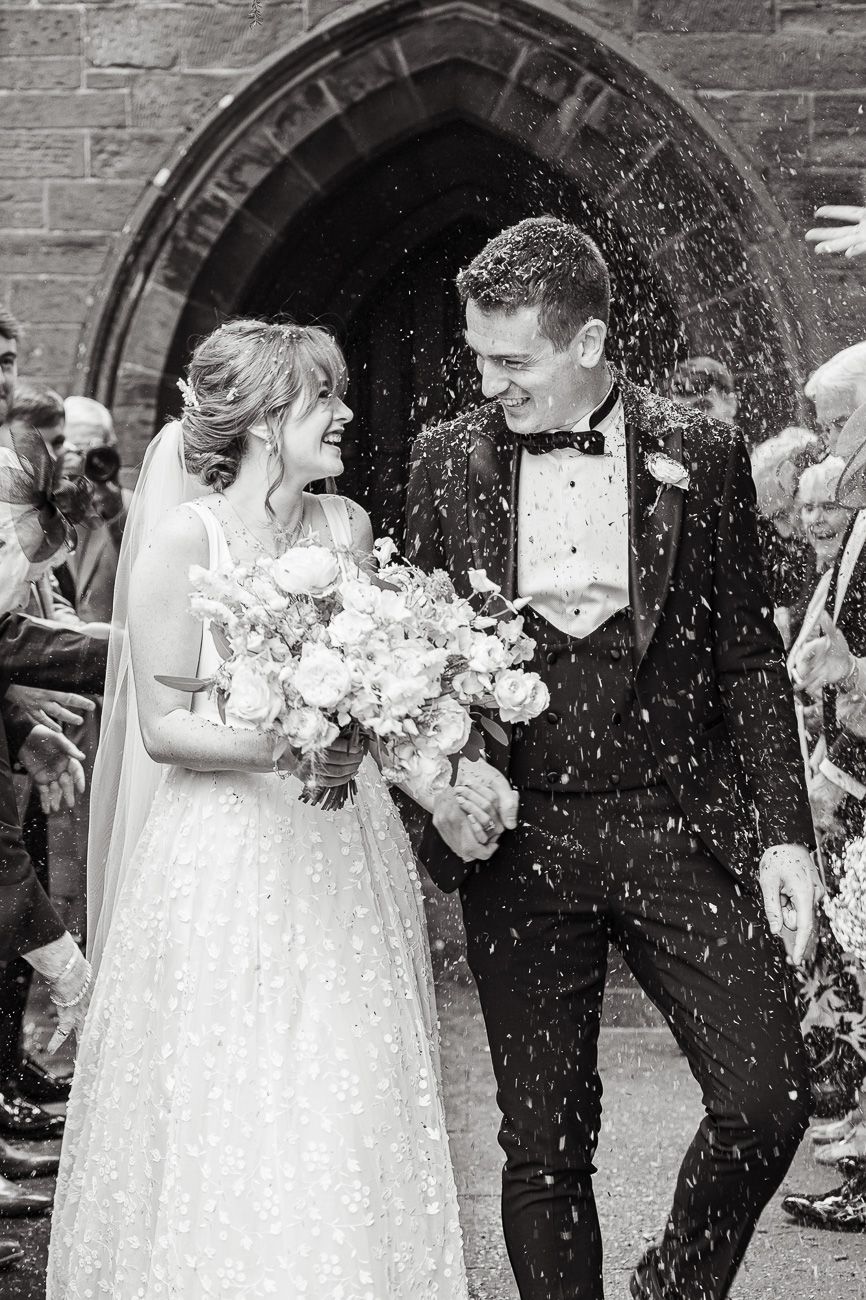 Black and white photo of a happy newlywed couple exiting church as guests shower them with rice after wedding ceremony.