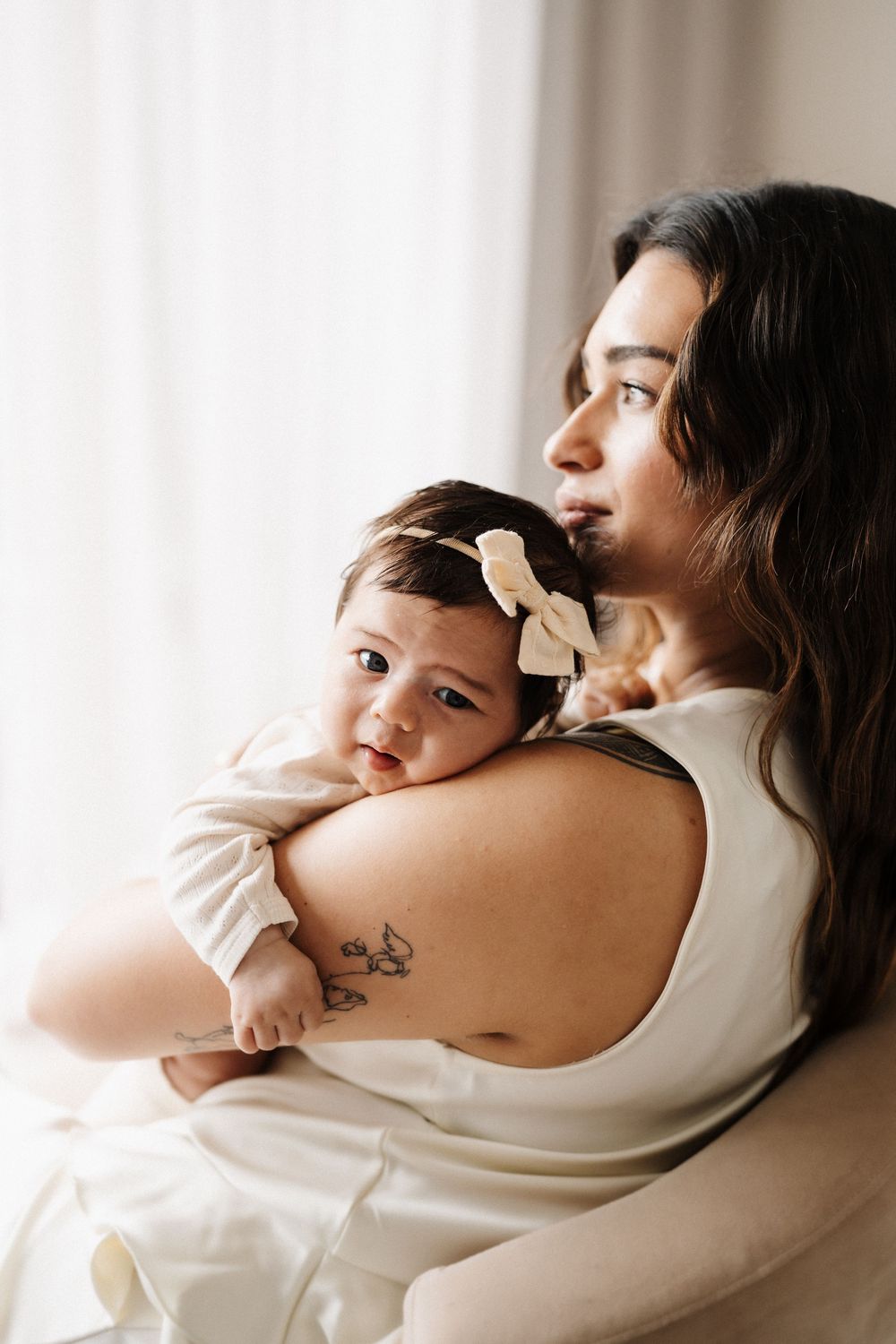 Parent in white tank top holding sleeping infant with bow near bright window.