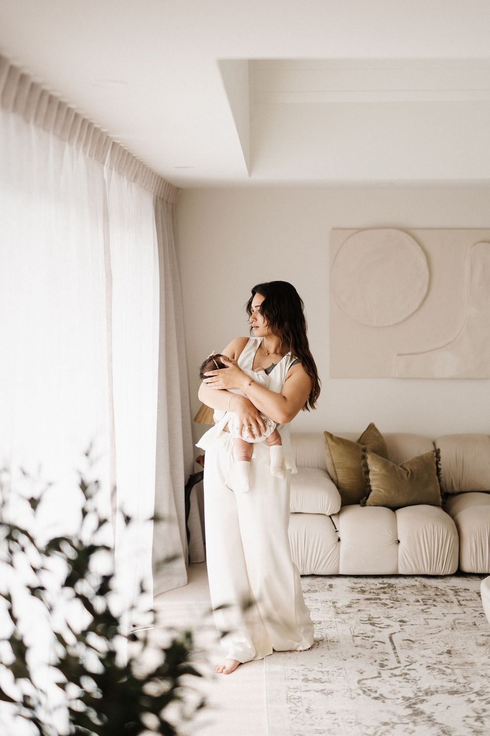 Parent in white outfit holding infant in modern living room with minimal decor.