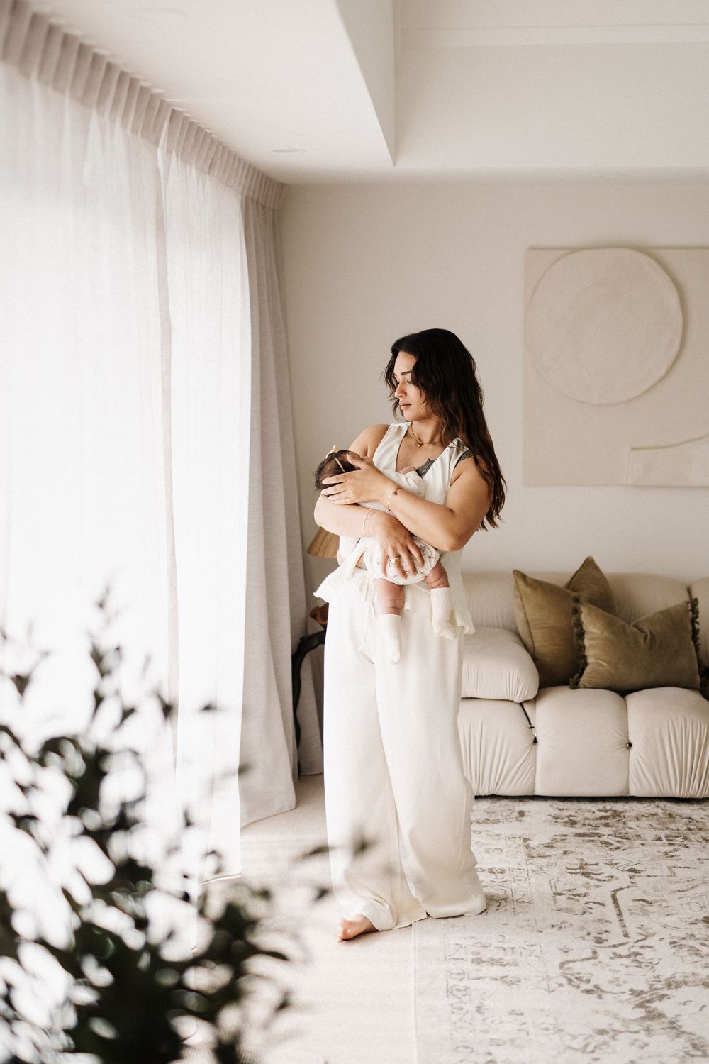 A mother in white clothing holds her baby while standing in a minimalist living room with natural light.