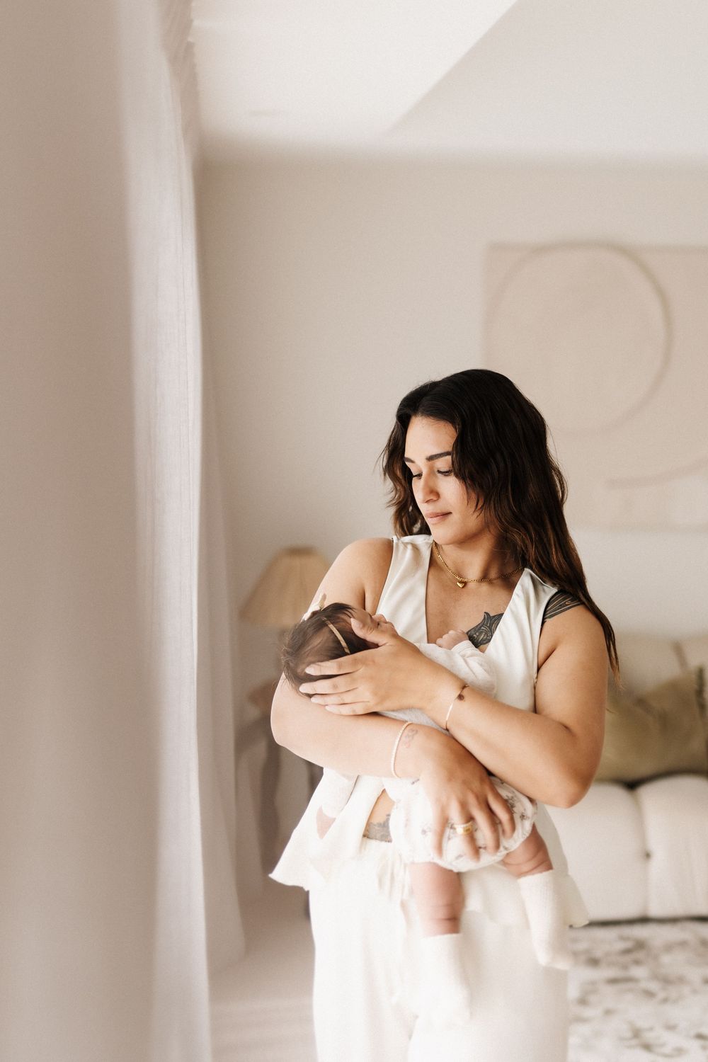 A woman in a white dress holds a newborn baby tenderly in a bright, minimalist room with neutral decor.