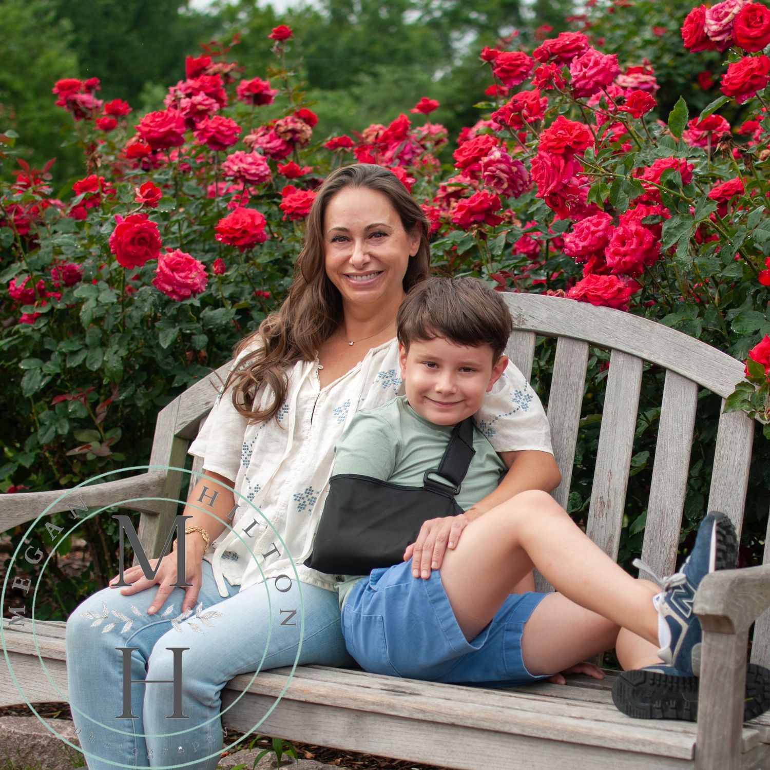 A mother and son sitting together on a wooden garden bench surrounded by blooming red rose bushes on a summer day.