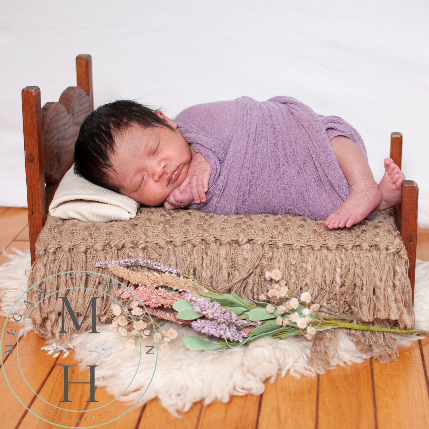 Sleeping baby rests on a rustic wooden bed with beige textured bedding and delicate flowers on hardwood flooring.