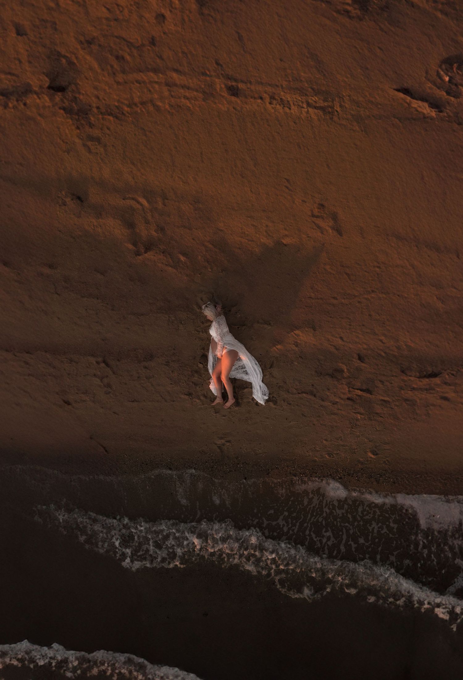 Aerial view of grey dog sitting on red rock cliff edge overlooking dark ocean waves below at Half Moon Bay, Black Rock, Victoria