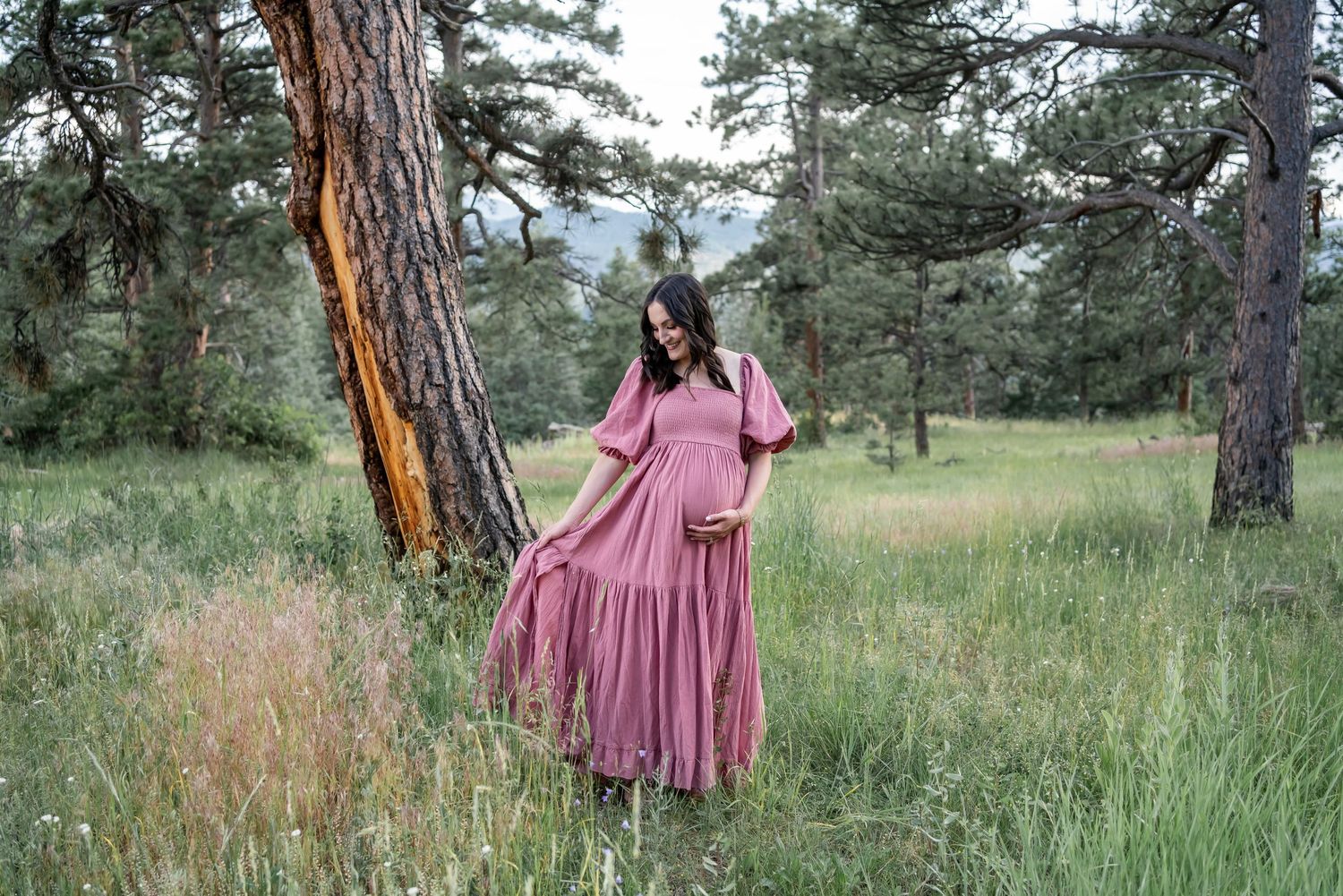 Someone in a flowing mauve maxi dress stands gracefully among tall grasses and pine trees in a natural outdoor setting.