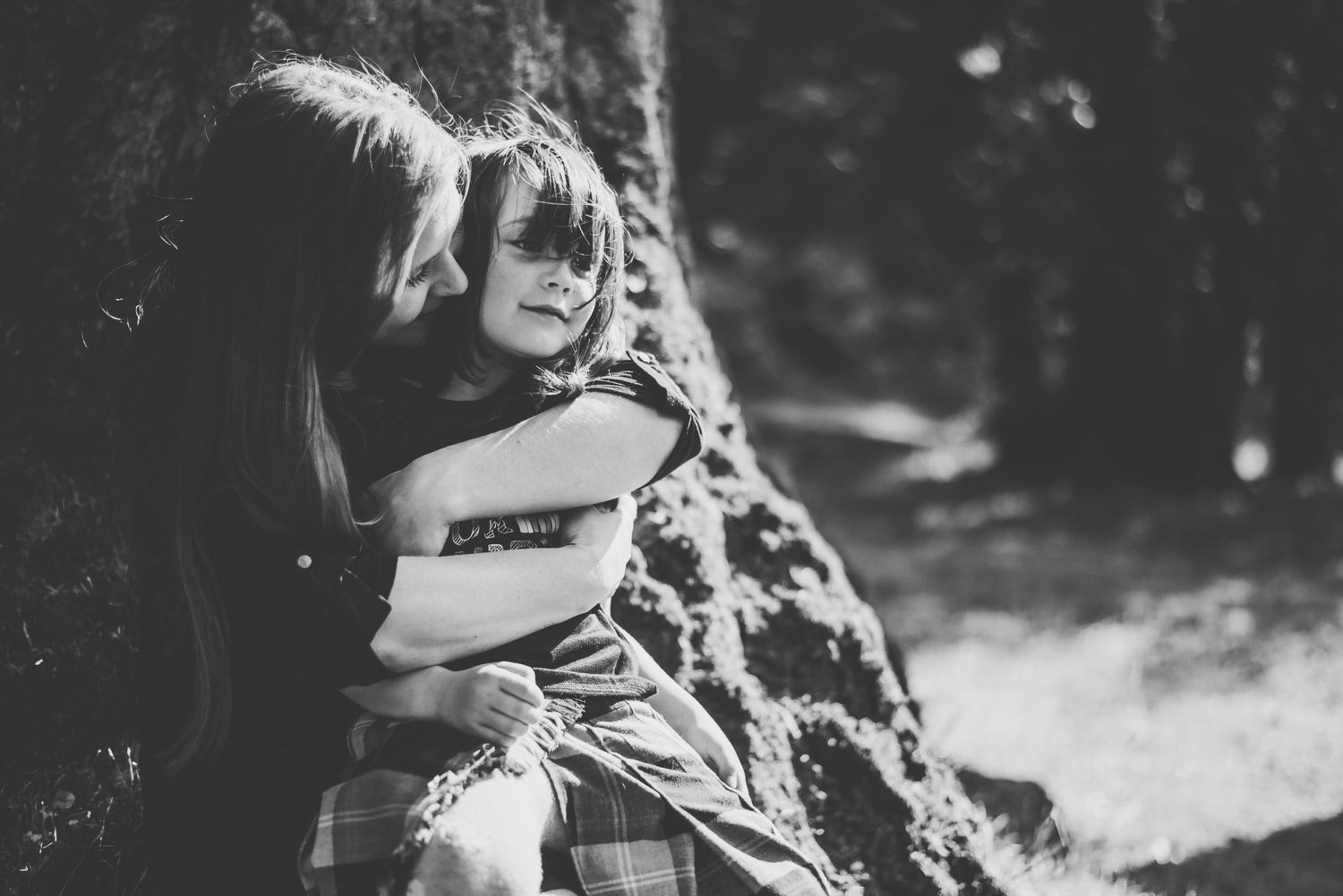 A tender moment as a mother embraces her child, both sitting peacefully against a tree in the soft outdoor light.