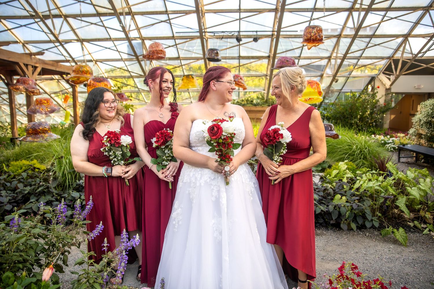 Wedding party wearing burgundy dresses and white gown pose together in greenhouse with flowers and greenery.