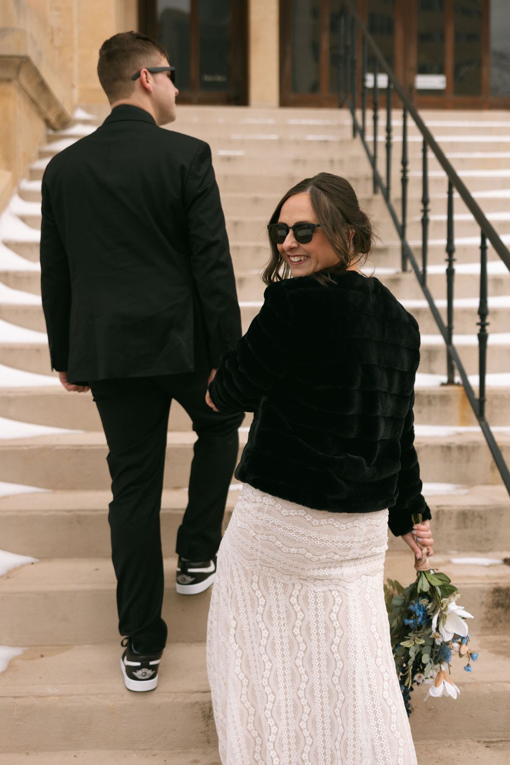 Two people in formal attire walk up stone steps, with one holding a bouquet while wearing a black top and white skirt.