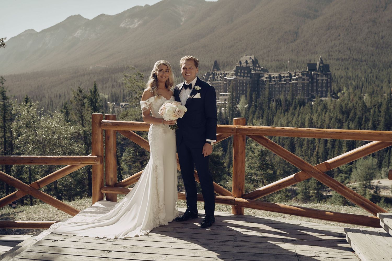 A wedding couple poses on a wooden deck overlooking the majestic Banff Springs Hotel nestled in the Canadian Rockies. Wedding photography