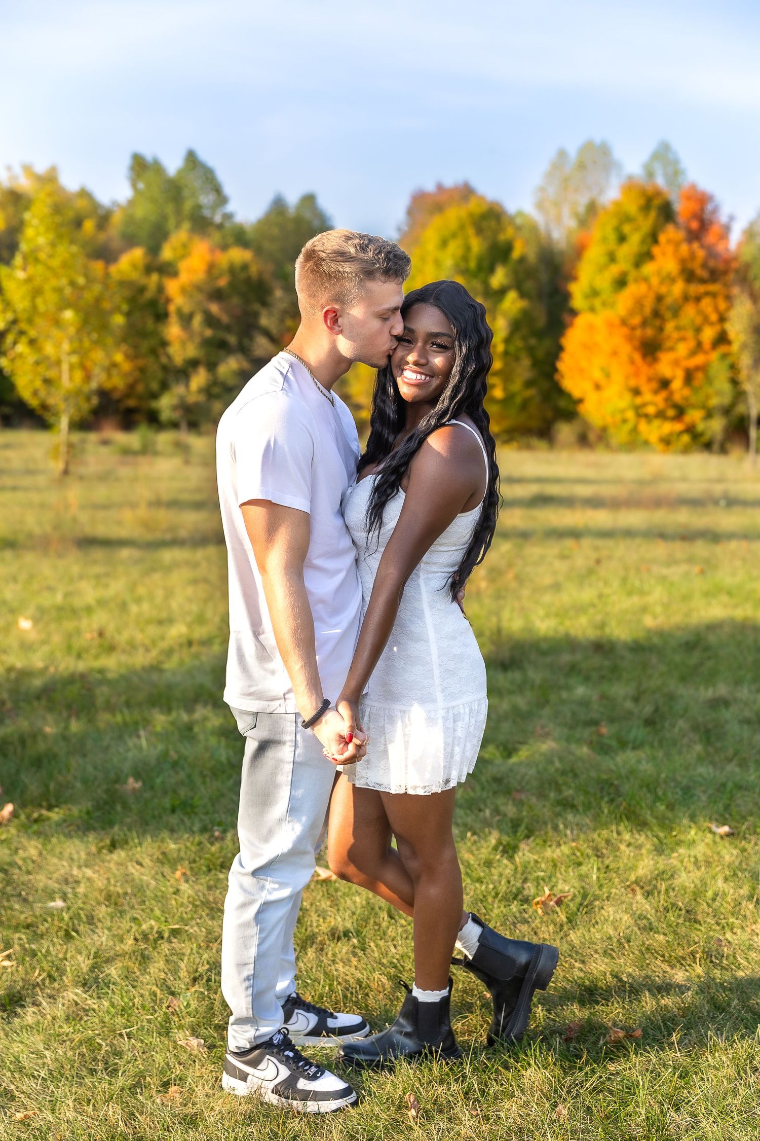 A couple embraces in a field during autumn with colorful fall foliage trees in the background.