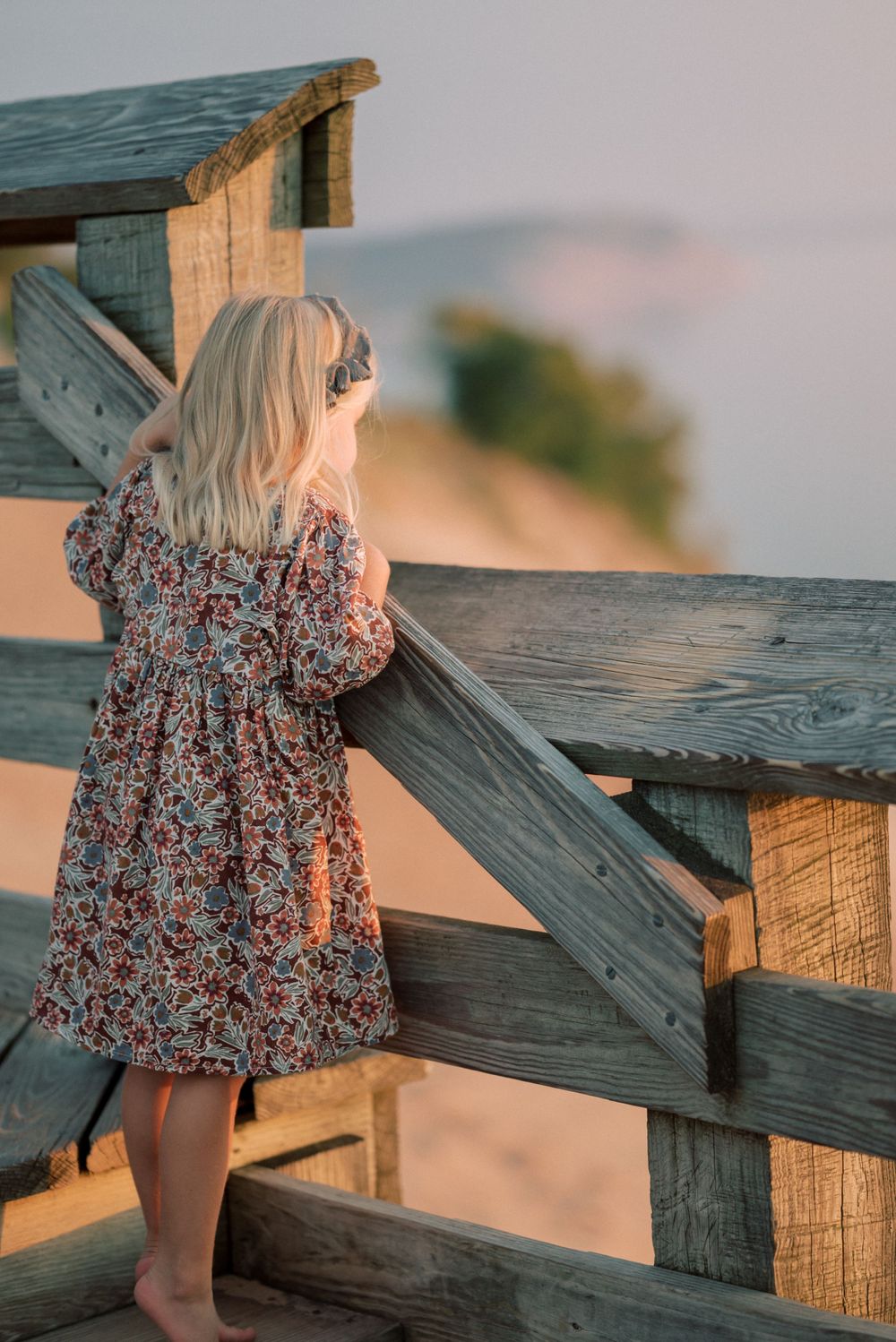 Child in floral dress looking out from wooden deck railing at sunset.