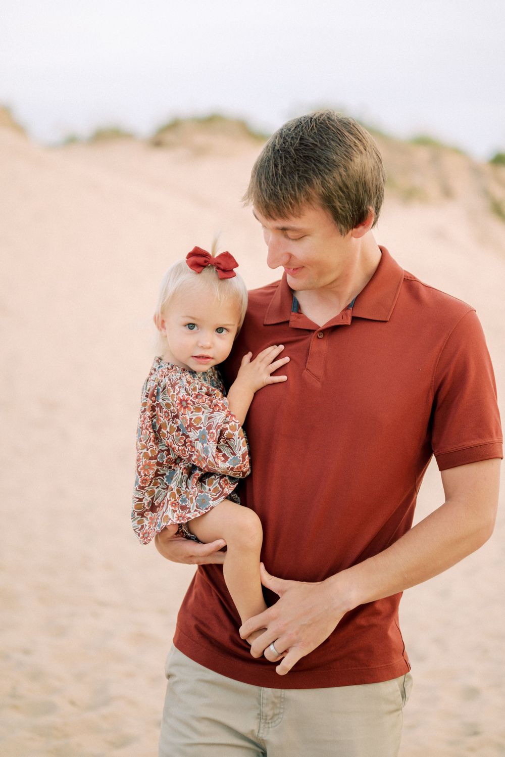 Parent in red polo shirt holding young child wearing floral dress on sandy dunes.